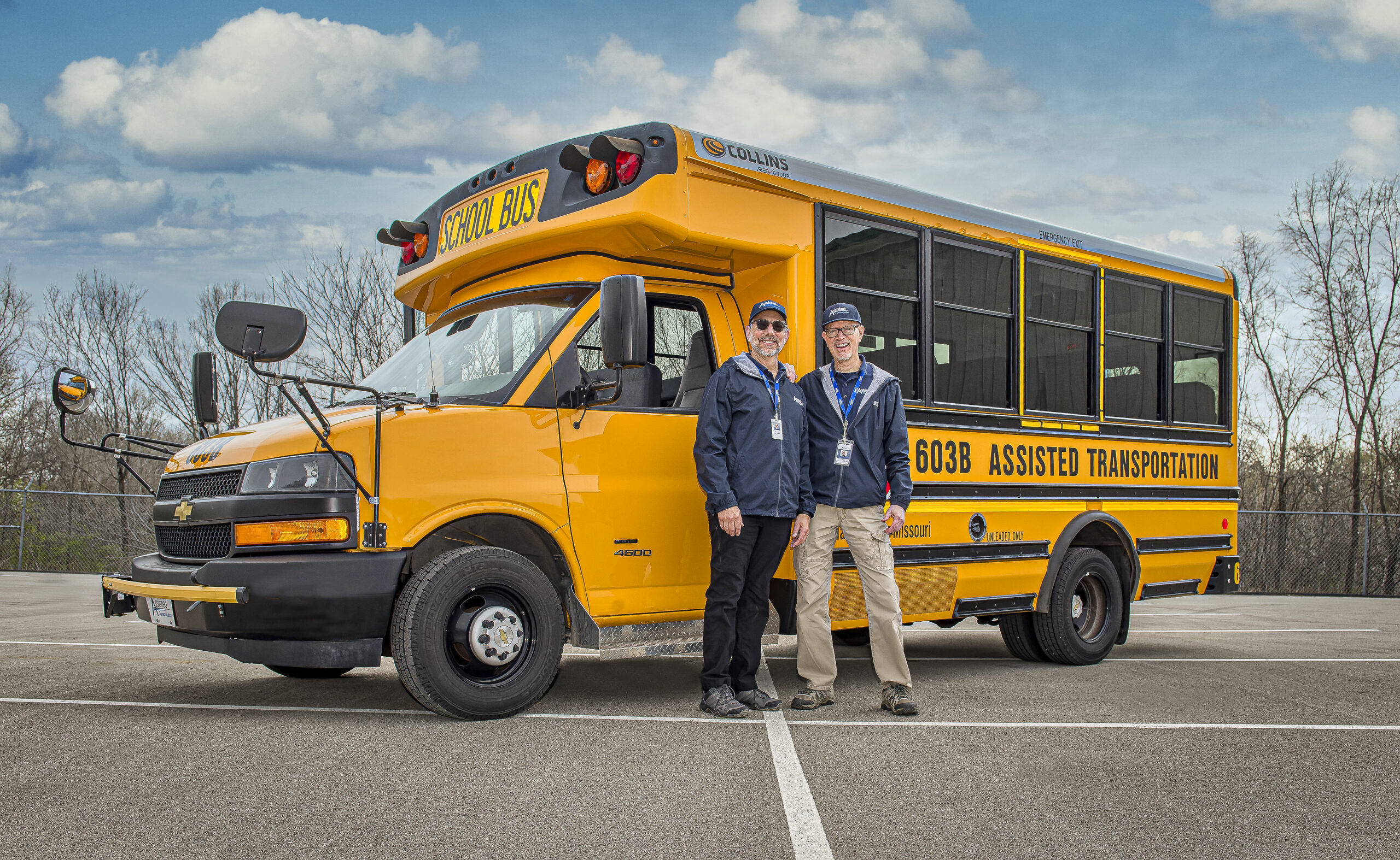 2 Assisted Drivers stand in front of a small yellow school bus.